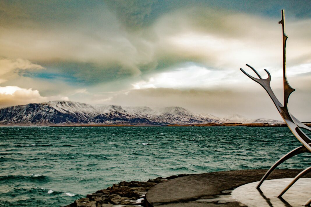 islandia Dramatyczny islandzki krajobraz z oceanem, górami i nowoczesną rzeźbą pod tętniącym życiem niebem.