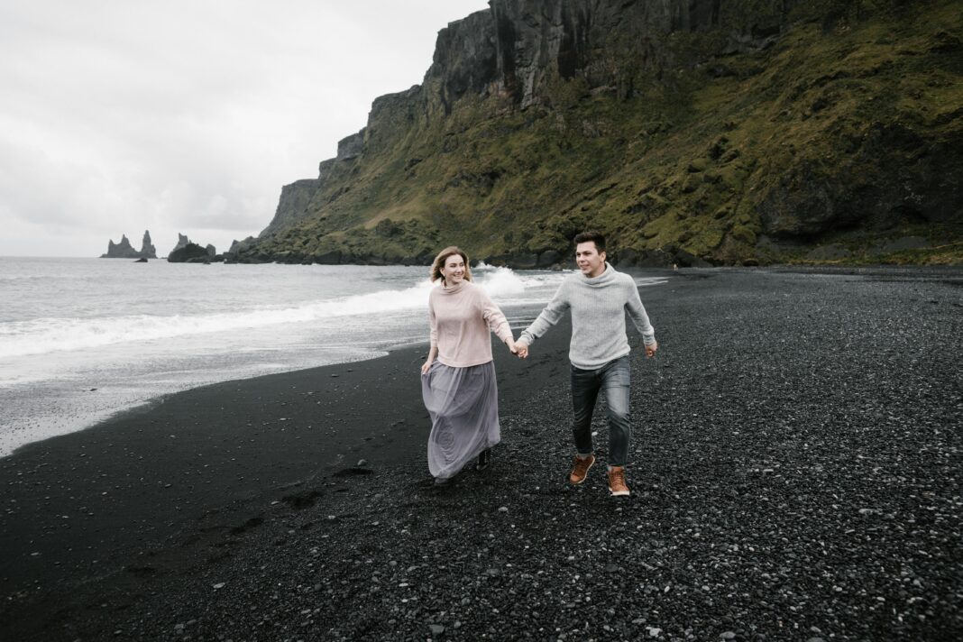 islandia Szczęśliwa kochająca para trzymająca się za ręce i odkrywająca wspaniałą czarną żwirową plażę, ciesząc się weekendem na klifowym wybrzeżu