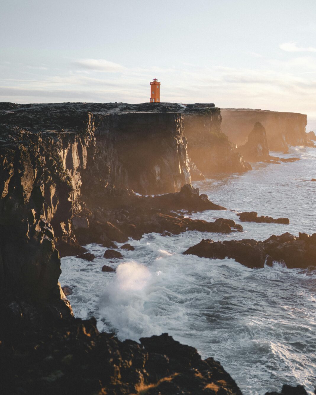 islandia Oszałamiający widok na latarnię morską na nierównym wybrzeżu Islandii podczas zachodu słońca, ukazujący spektakularne klify i fale oceanu.