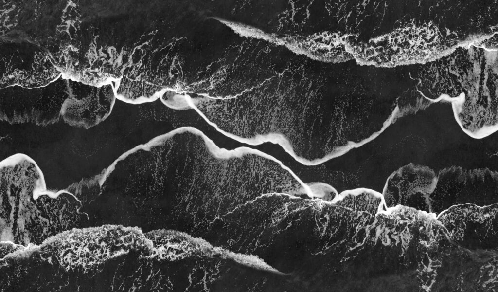 Oszałamiające monochromatyczne ujęcie lotnicze fal oceanicznych rozbijających się o brzeg, tworząc abstrakcyjne wzory.