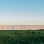 Spokojny widok na Jezioro Tyberiadzkie z trawiastym pierwszym planem i odległymi wzgórzami oświetlonymi wschodem słońca.