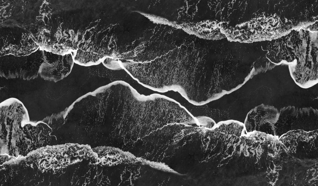 Oszałamiające monochromatyczne ujęcie lotnicze fal oceanicznych rozbijających się o brzeg, tworząc abstrakcyjne wzory.