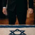 Mężczyzna w garniturze stoi za stołem z powieszoną izraelską flagą.