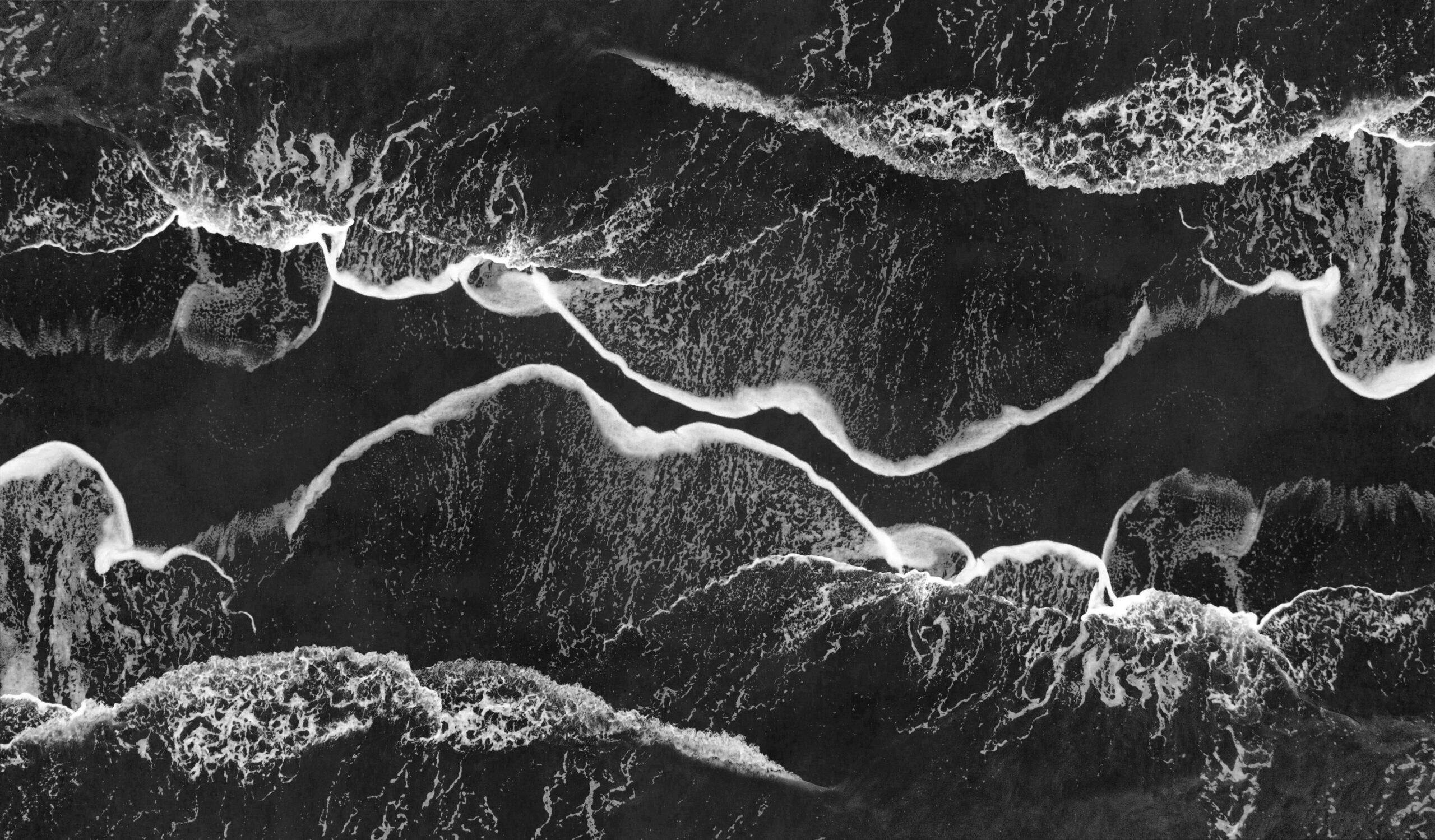 Oszałamiające monochromatyczne ujęcie lotnicze fal oceanicznych rozbijających się o brzeg, tworząc abstrakcyjne wzory.