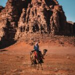 Mężczyzna jedzie na wielbłądzie po malowniczej pustyni Wadi Musa w Jordanii, gdzie widać spektakularne formacje skalne.