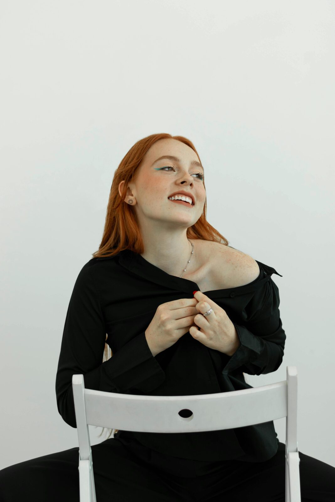 Portret wesołej rudowłosej kobiety w czarnym stroju, siedzącej na krześle i figlarnie pozującej w pomieszczeniu.