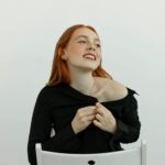 Portret wesołej rudowłosej kobiety w czarnym stroju, siedzącej na krześle i figlarnie pozującej w pomieszczeniu.