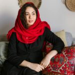 Petrografy z Eshkiolmes: Ślady dawnych cywilizacji Kobieta w czerwonym szaliku i czarnej sukience siedzi w pomieszczeniu, prezentując tradycyjny styl.