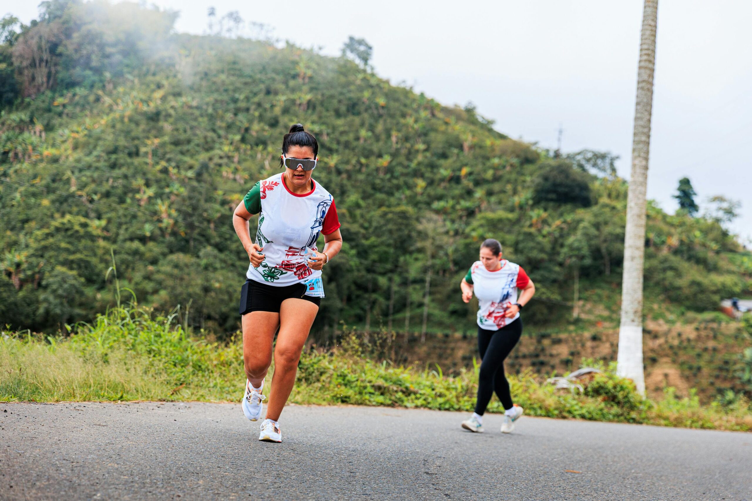 Dwie kobiety biegające po bujnym, pagórkowatym krajobrazie Pereira w Risaraldzie w Kolumbii, prezentując ćwiczenia na świeżym powietrzu.