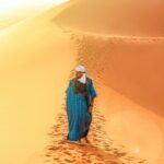 Osoba ubrana w tradycyjny strój spaceruje po wydmach Sahary o wschodzie słońca.