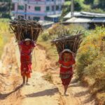 Dwie kobiety w tradycyjnym stroju niosące drewno opałowe w wiejskim nepalskim krajobrazie.