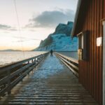 Norwegia – jak zaplanować budżetową podróż? Spokojne ujęcie zaśnieżonego molo w Norwegii o zachodzie słońca z samotną postacią wpatrującą się w morze.