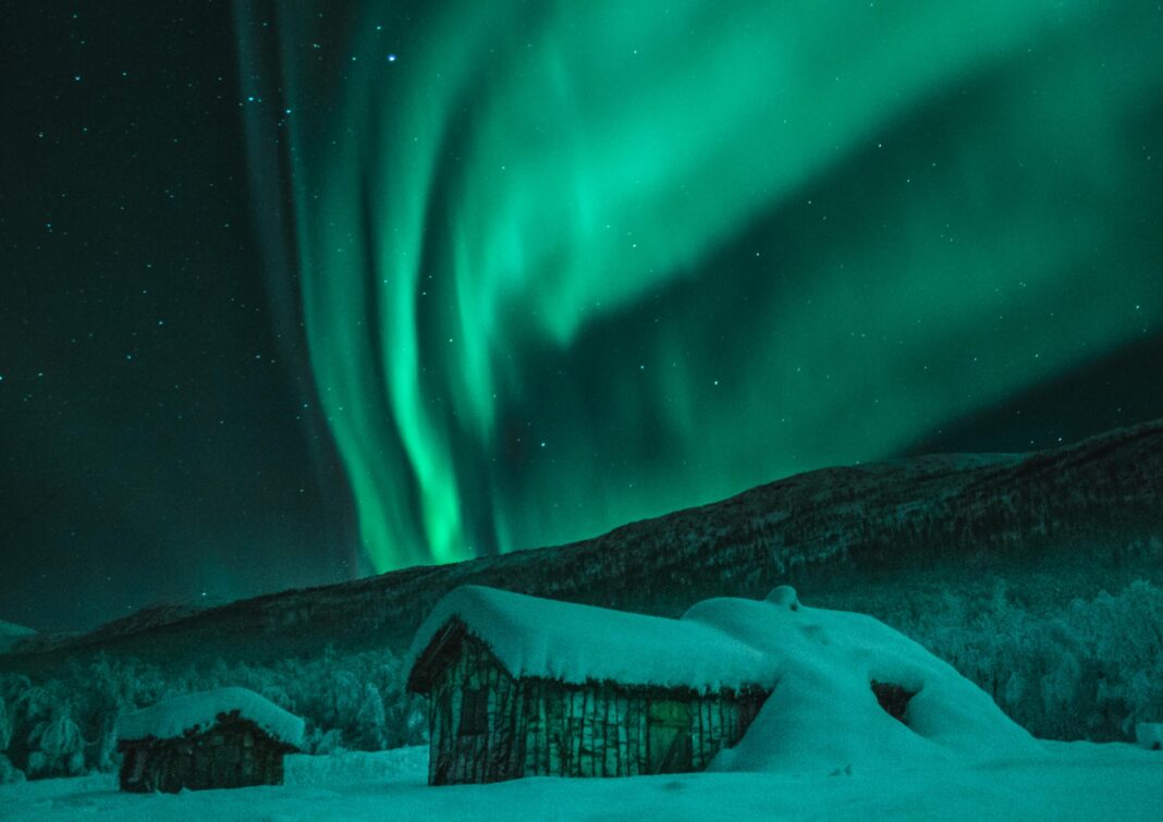 norwegia Urzekający widok zorzy polarnej oświetlającej zaśnieżone kabiny i zimowe nocne niebo w Norwegii.