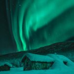Zorza polarna w Norwegii – gdzie i kiedy ją zobaczyć Urzekający widok zorzy polarnej oświetlającej zaśnieżone kabiny i zimowe nocne niebo w Norwegii.