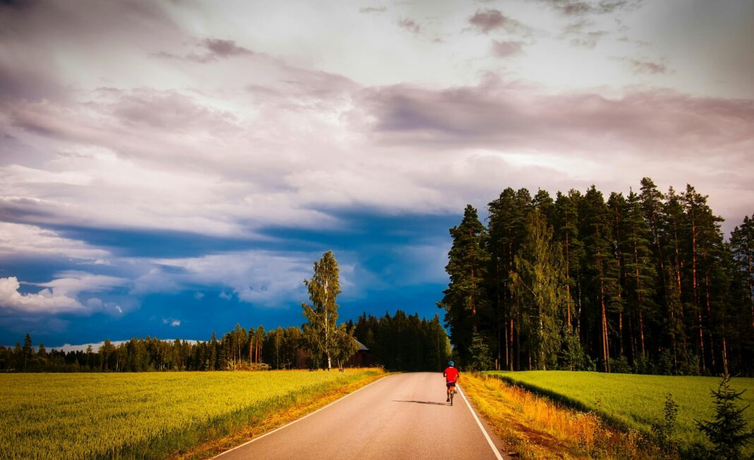 podroz Samotny rowerzysta pedałuje wiejską drogą otoczoną bujnymi polami i lasami pod dramatycznie zachmurzonym niebem.