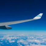 Widok skrzydła samolotu lecącego wysoko nad chmurami na tle czystego, błękitnego nieba.