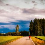 Samotny rowerzysta pedałuje wiejską drogą otoczoną bujnymi polami i lasami pod dramatycznie zachmurzonym niebem.