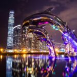 Dramatyczny pejzaż miejski nocą z oświetloną rzeźbą delfinów z odbiciami.