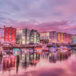 Fakty i mity o tanich lotach do Lizbony Tętniąca życiem scena na nabrzeżu z kolorowymi budynkami i łodziami odbijającymi się w wodzie o zachodzie słońca.