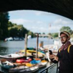 Mężczyzna w kapeluszu i szaliku korzystający ze smartfona nad malowniczą rzeką z łodziami w słoneczny dzień.