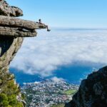 Osoba siedzi na krawędzi klifu, podziwiając zapierający dech w piersiach widok na ocean i miasto poniżej.