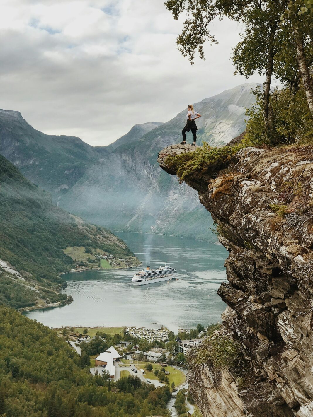 Osoba cieszy się zapierającym dech w piersiach widokiem z klifu nad fiordem Geiranger w Norwegii, z czystym niebem i statkiem wycieczkowym poniżej.