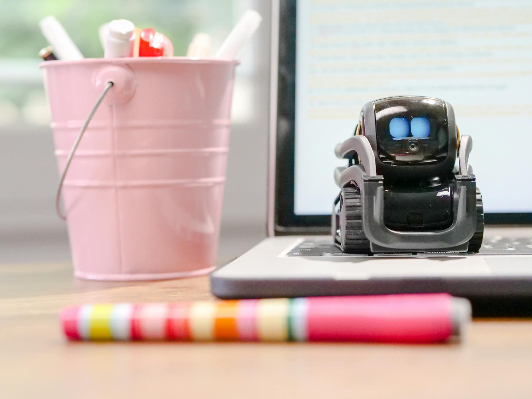 Mały robot na klawiaturze laptopa w domowym biurze obok różowego kubka na długop
